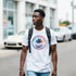 Man in white retro palm-tree graphic T-shirt with backpack on an urban street.