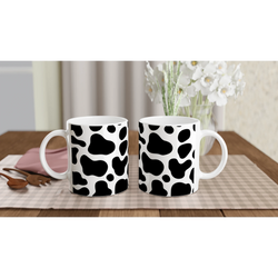 Three black-and-white cow-print mugs on a wooden table with a vase of white flowers in the background.