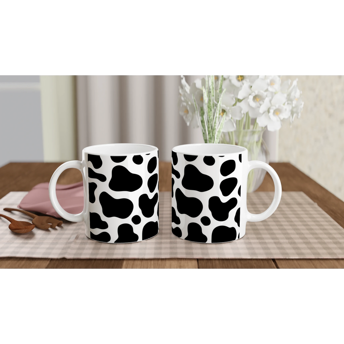 Three black-and-white cow-print mugs on a wooden table with a vase of white flowers in the background.