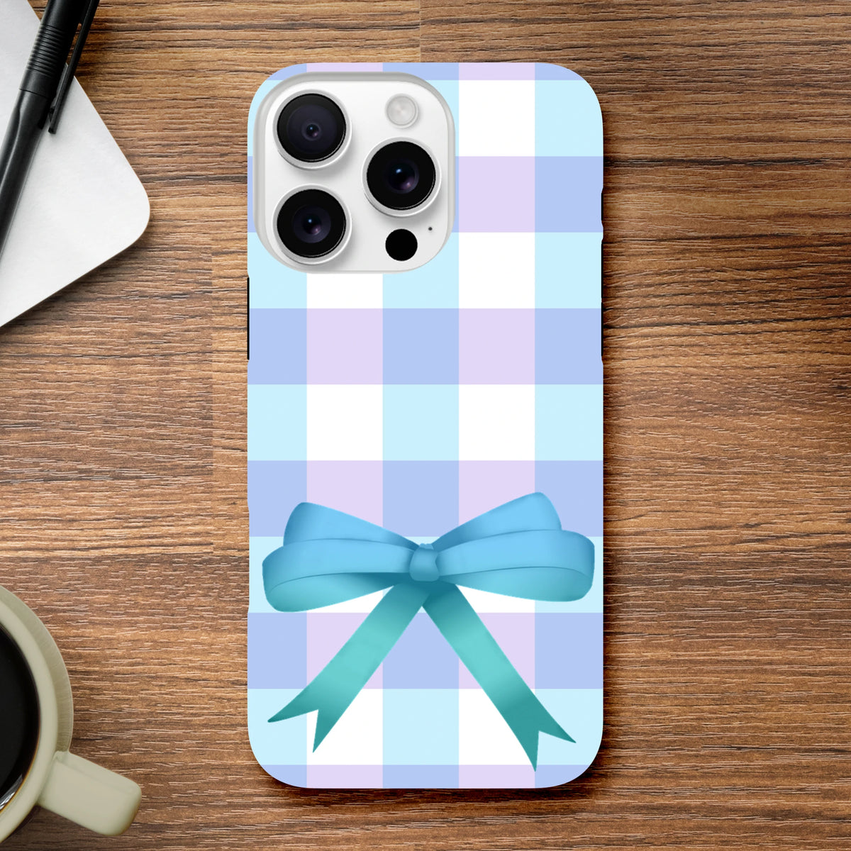 Blue gingham iPhone case with a 3D teal bow, featuring soft lavender and white checks on a wooden desk.