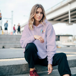 Woman wearing a lavender hoodie with a colorful circular palm tree chest logo, crouching on concrete steps in an urban setting.