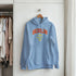 Light blue Berlin graphic women’s hoodie with red lettering and yellow accents on a hanger in a closet.