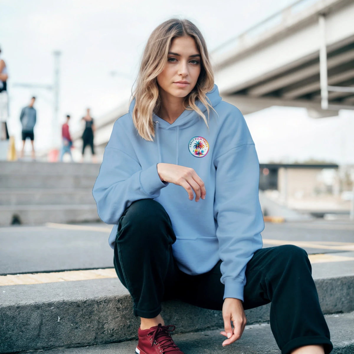Woman wearing light blue tropical hoodie with palm tree graphic, styled with black pants and red sneakers for a laid-back urban look.