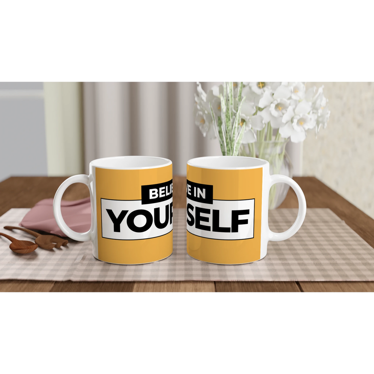 Pair of yellow Believe Yourself mugs with bold slogan text on a wooden table near a vase of white flowers.