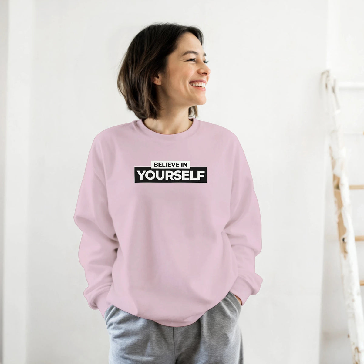 Woman wearing pastel pink Believe In Yourself sweatshirt with bold black slogan.