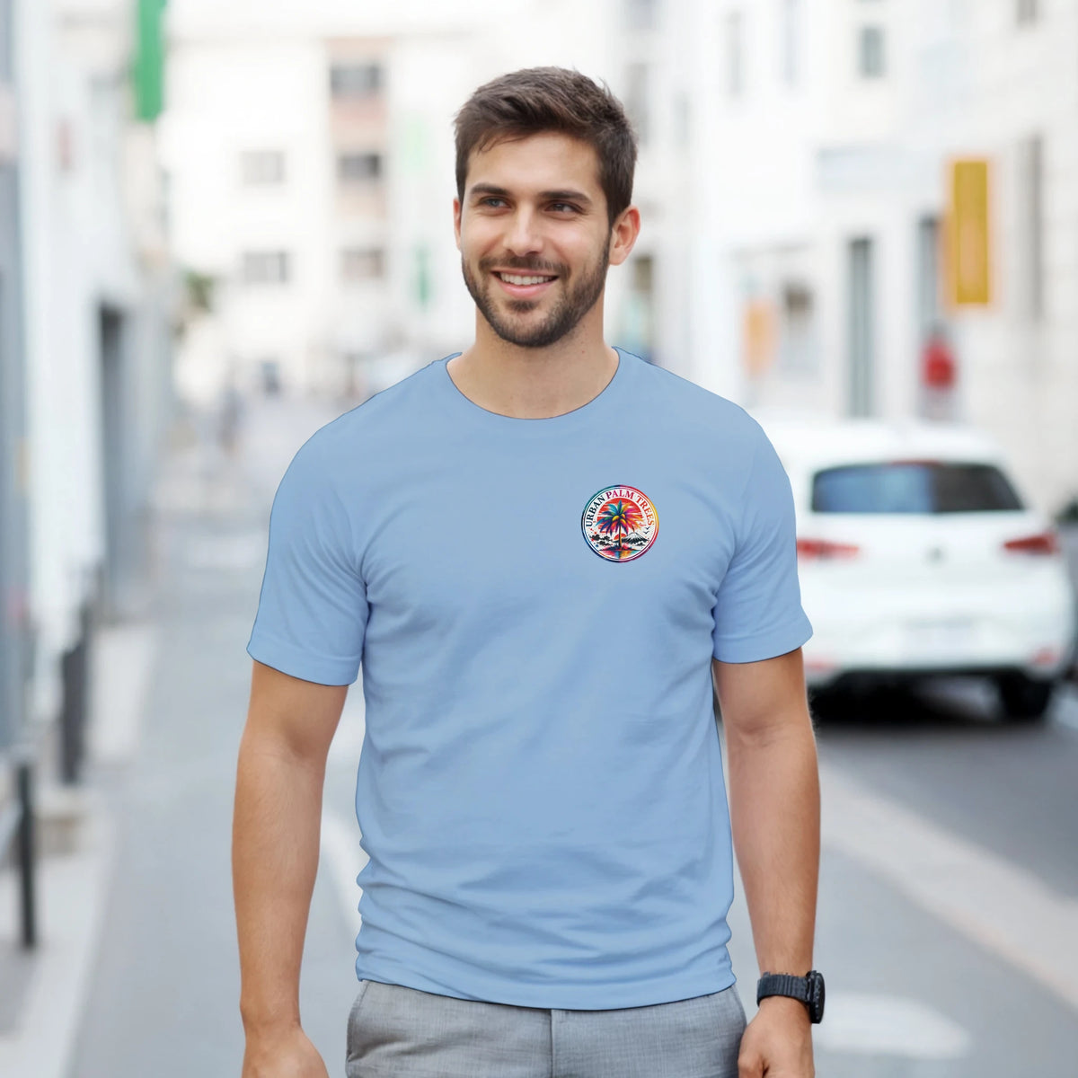 Man wearing light blue t-shirt with small circular Urban Palm Trees graphic on chest, standing on a city street.