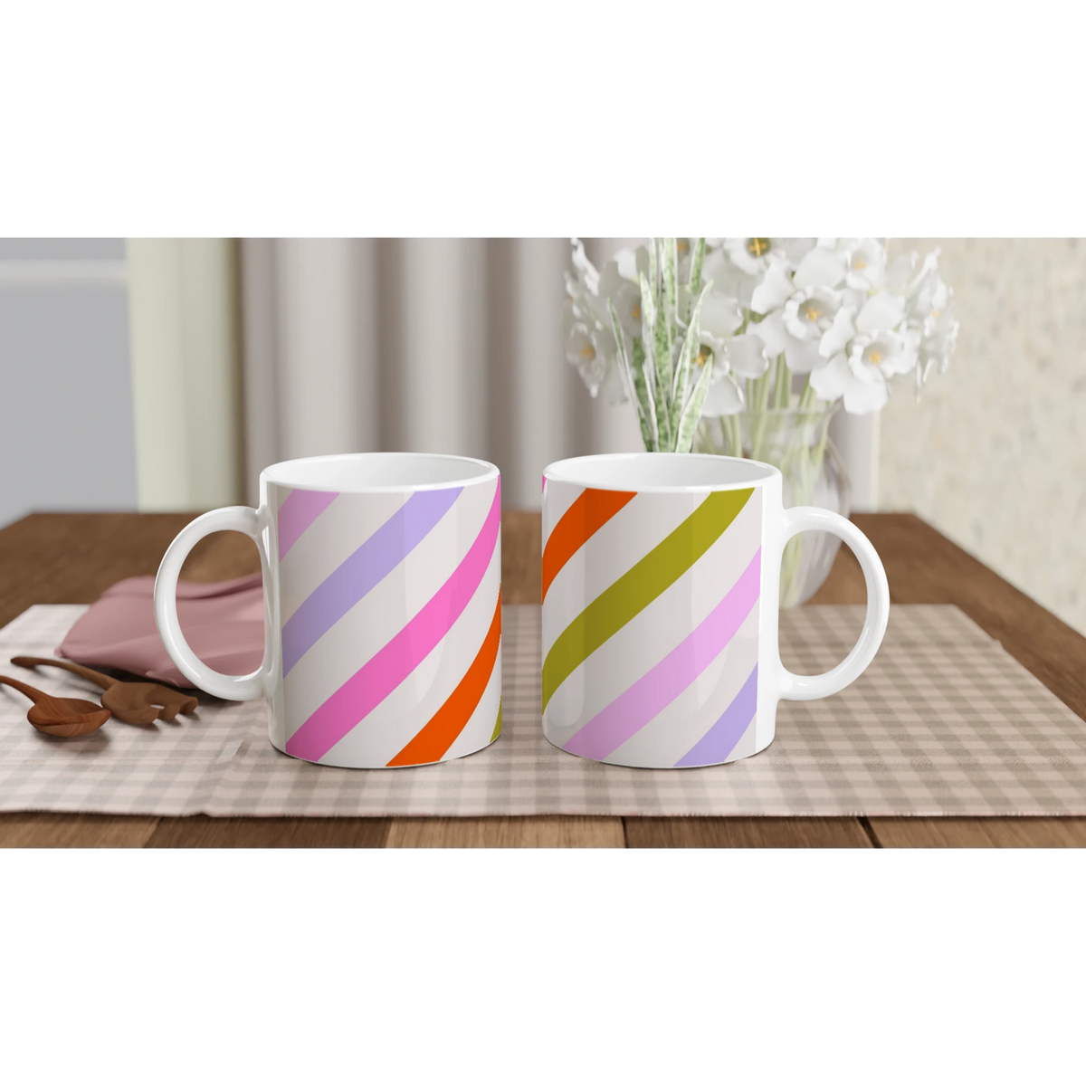 Pastel diagonal stripe ceramic mugs with rainbow diagonals on white, set on a table near a vase of white flowers.