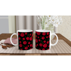 Two white ceramic mugs featuring a vibrant red cherry pattern on a black background, set on a checkered table mat with wooden spoons and white flowers.