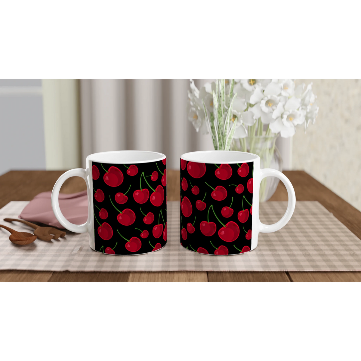 Two white ceramic mugs featuring a vibrant red cherry pattern on a black background, set on a checkered table mat with wooden spoons and white flowers.