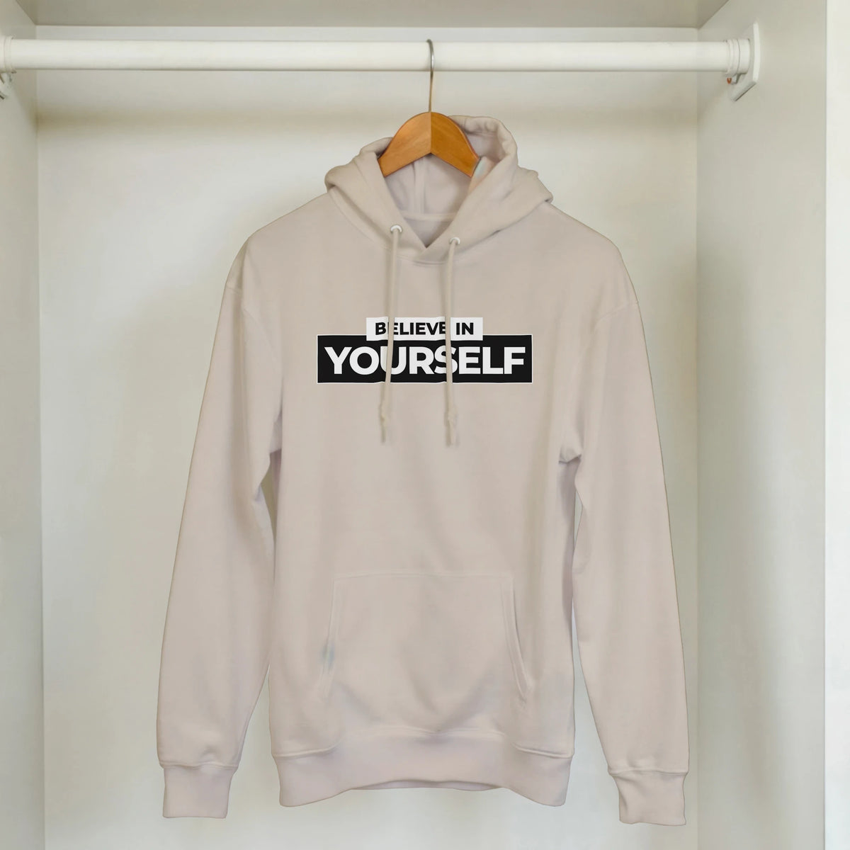 Beige women's Believe In Yourself hoodie on a wooden hanger in a closet.