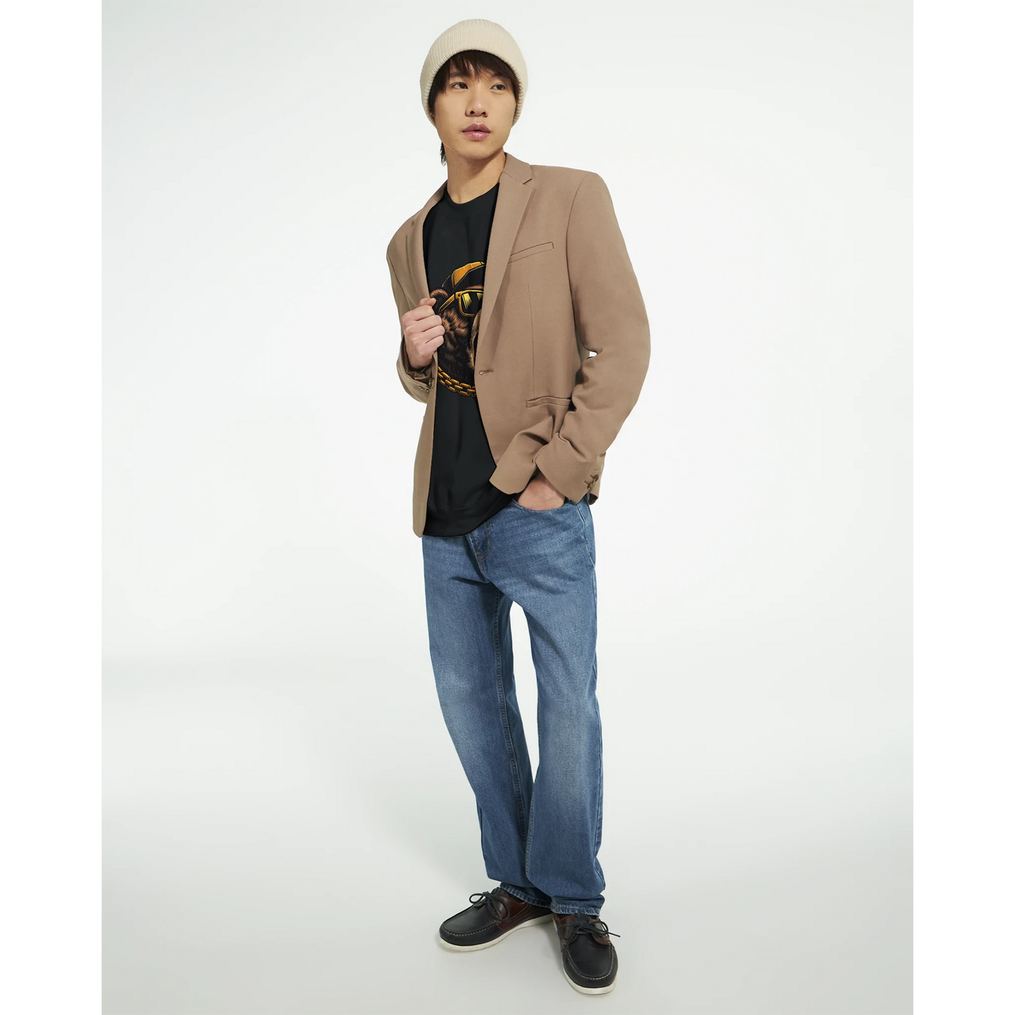 Person wearing a beige blazer, black men's graphic sweatshirt, blue jeans, and a beige cap on a white background