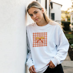Woman in light gray sweatshirt featuring an orange gingham bow graphic, leaning against a wall outdoors.