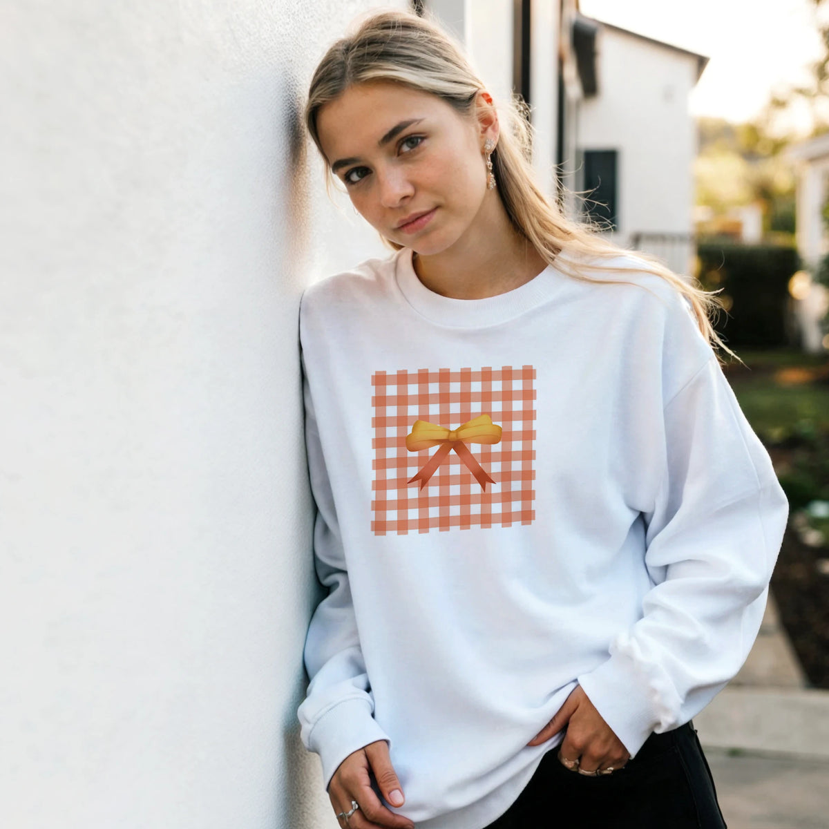 Woman in light gray sweatshirt featuring an orange gingham bow graphic, leaning against a wall outdoors.