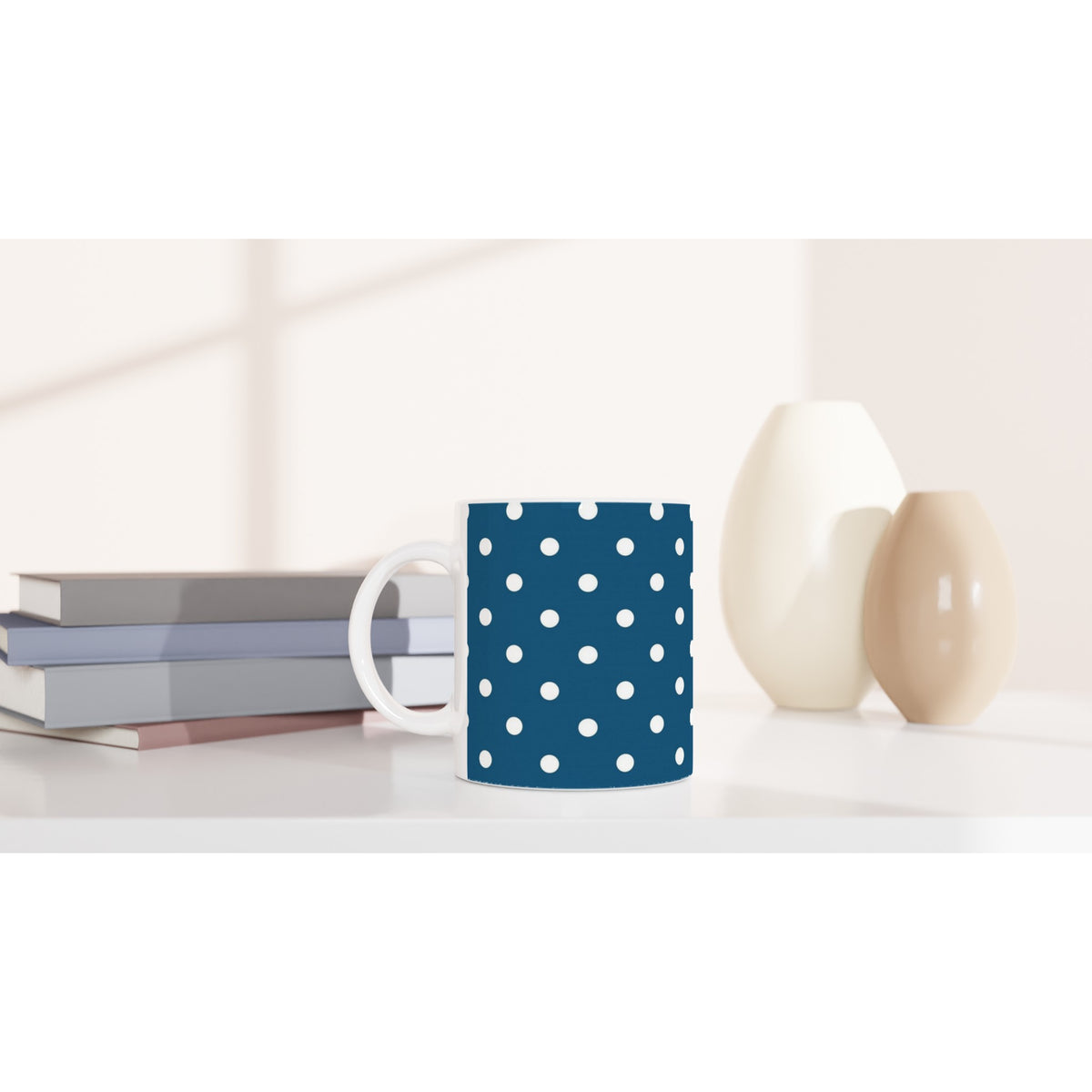 Navy blue ceramic mug with white polka dots sits on a white desk next to stacked books and minimalist vases.