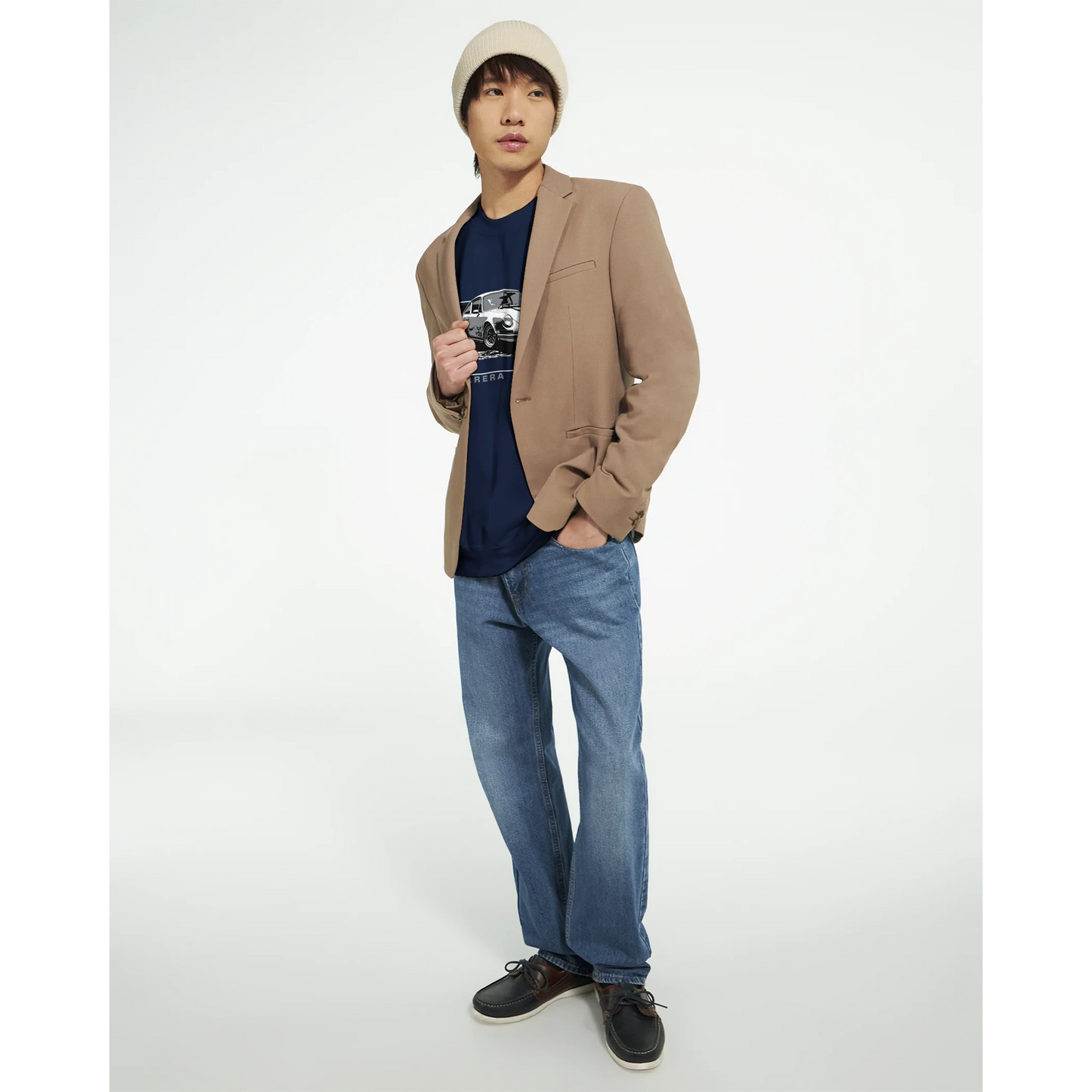 Male model in a camel blazer over a navy car-graphic tee, blue jeans, beanie, and dark loafers in a studio.