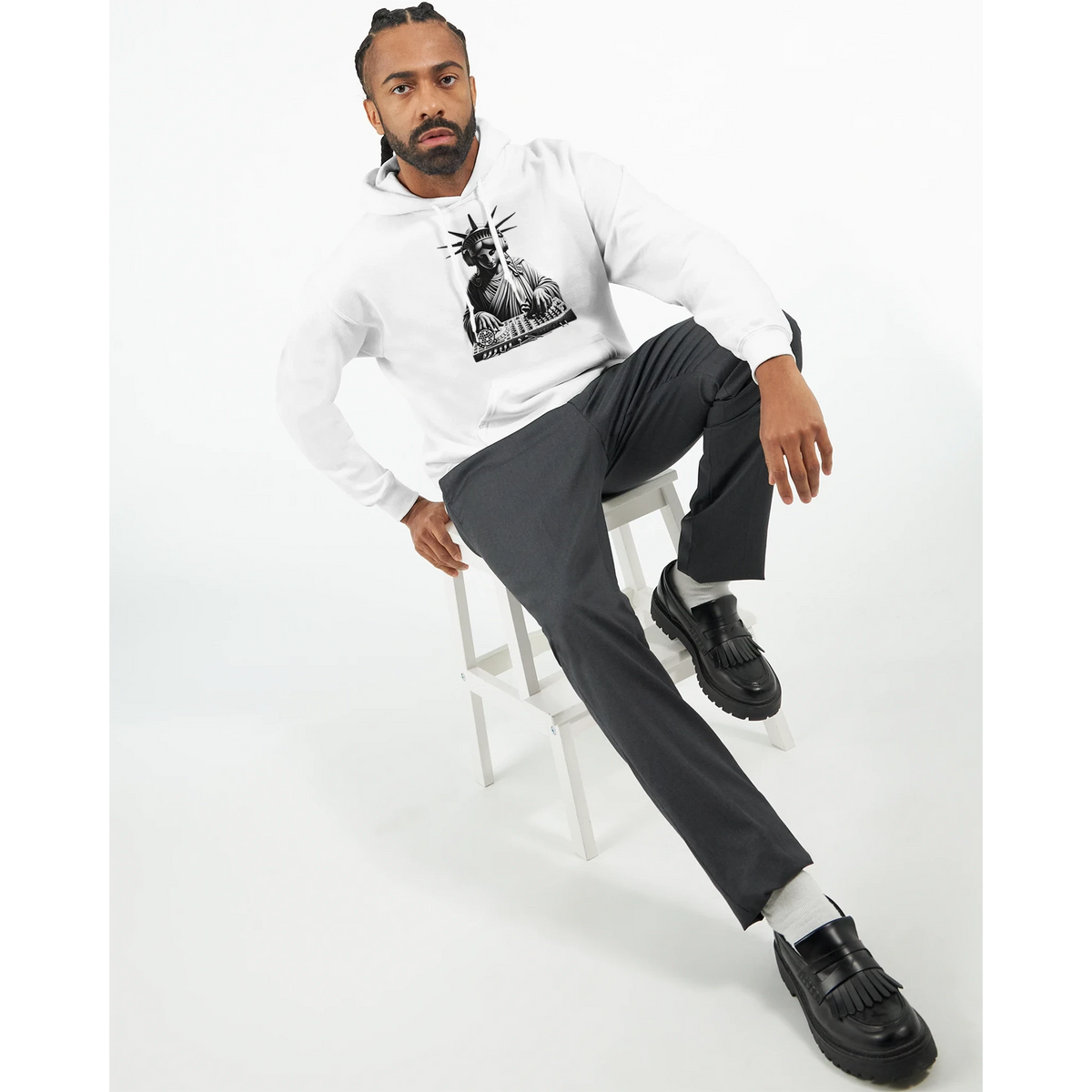 Man wearing Liberty DJ graphic hoodie in white, seated on a stool with grey pants and black loafers.