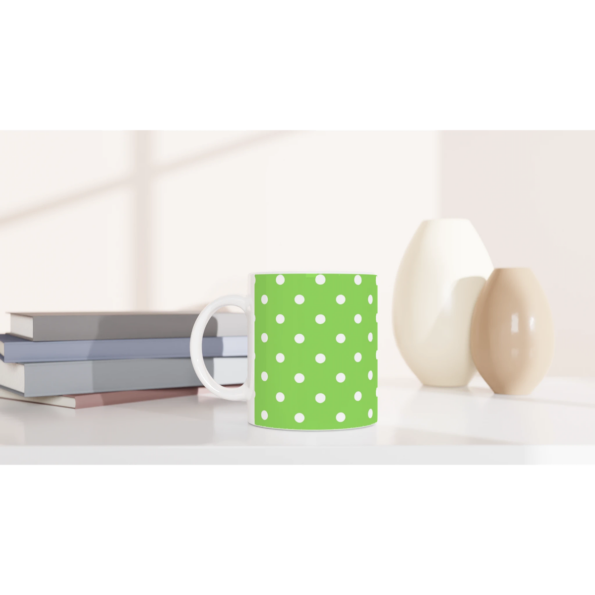 Green and white polka dot ceramic mug sitting on a white desk next to stacked books and minimalist vases.