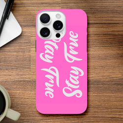 Pink iPhone case featuring vertical "Stay True" white script text, designed as a positive quote cover for iPhone 13 through 16 models on a wooden desk.