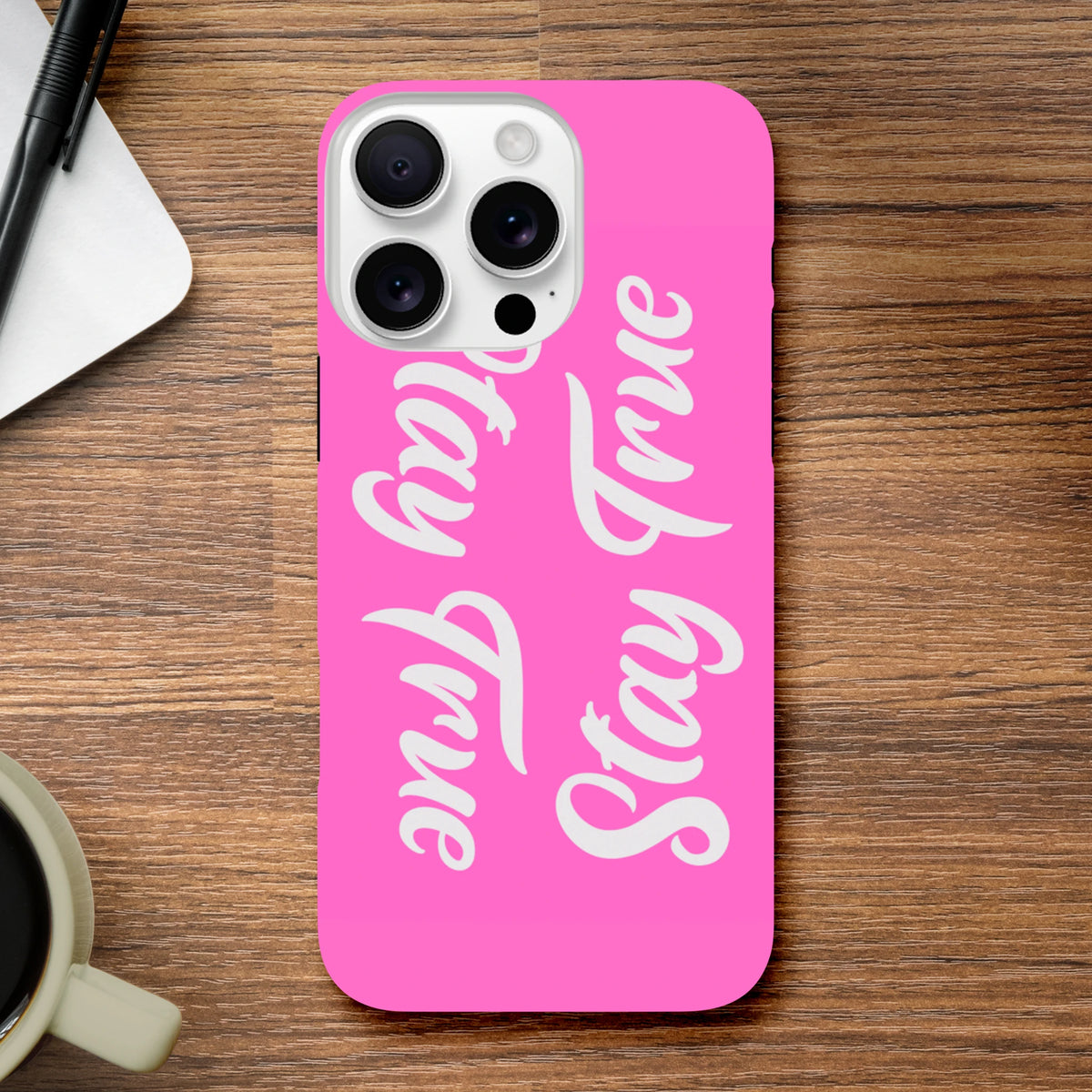 Pink iPhone case featuring vertical "Stay True" white script text, designed as a positive quote cover for iPhone 13 through 16 models on a wooden desk.