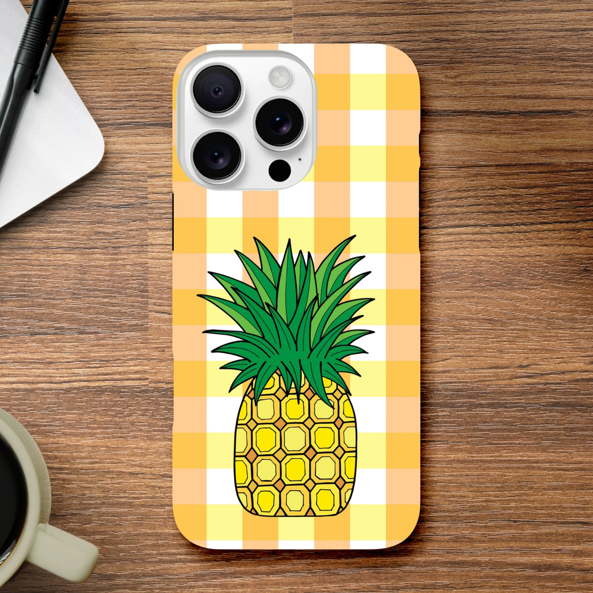Yellow gingham iPhone case featuring a cute tropical pineapple illustration on a wooden desk surface.
