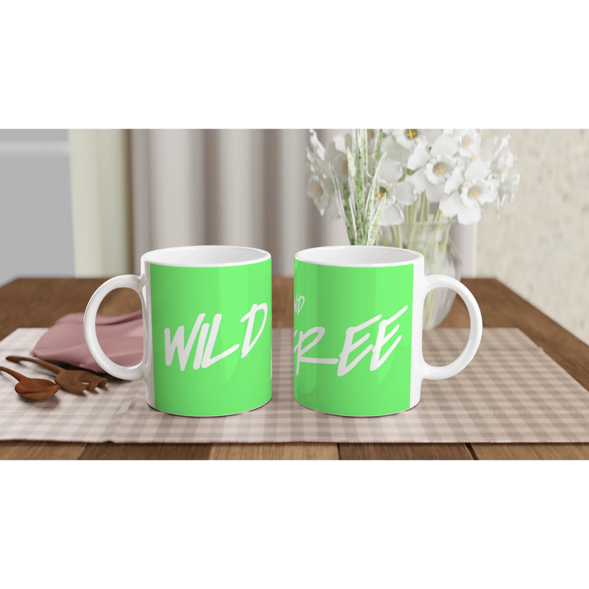 Two green mugs with white cursive slogan on a wooden table, bouquet in blurred background.