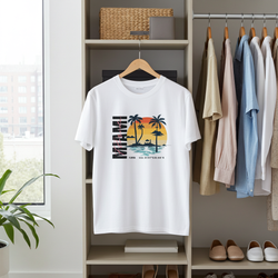 White t-shirt with 'Miami' graphic design hanging on a rack in a room with shelves and plants.
