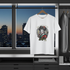White t-shirt with a graphic design hanging in front of a cityscape window.