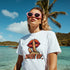White Lips graphic T-shirt on a woman, soft cotton blend with lips and finger over lips, 'SHUT UP' on the beach.