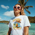 Woman wearing white Tropical Cocktail graphic T-shirt at the beach with pink sunglasses and palm trees.