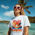 Woman wearing Hawaii Aloha graphic tee on white cotton-blend shirt at tropical beach with palm trees.