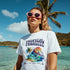 Woman in white Tropical Paradise graphic T-shirt with island scene and pink sunglasses on the beach.
