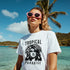 Model wearing white Tropical Paradise graphic T-shirt with pink sunglasses at a tropical beach.