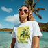 Woman wearing white Lemons graphic T-shirt with lemon print on a beach, pink sunglasses, tropical backdrop.