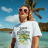 Woman in white lemon-graphic tee with lemon tree design on a tropical beach, pink heart-shaped sunglasses.