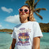 Woman wearing white soft cotton blend Mai Tai Dreams graphic tee by the beach with tropical sunset palm design.