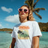 Woman wearing Tropical Dreamer tee with palm graphic on white soft cotton blend, beach scene, pink sunglasses.