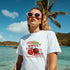 Woman wearing white Sweet Cherries graphic tee with red cherries at the beach.