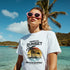 Woman in white Sunset Graphic T-Shirt with tropical palm sunset design on beach, pink sunglasses.