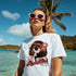 Woman wearing a white retro pop art graphic T-shirt at a sunny beach, pink heart-shaped sunglasses.