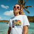 Woman wearing white Miami graphic T-shirt with palm trees and sunset by turquoise ocean, pink sunglasses.