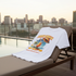 White t-shirt with a colorful graphic design on a lounge chair by a pool with city skyline in the background