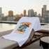White t-shirt with a colorful graphic design on a lounge chair by a pool with city skyline in the background