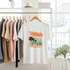 White t-shirt with 'Tropical Dreams' design hanging on a rack with other shirts.