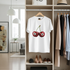 White t-shirt with cherry design on a hanger in a room with shelves and shoes.