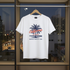 White t-shirt with palm tree design and 'California' text on a hanger in front of a cityscape.