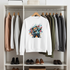 White sweatshirt with a colorful bear design hanging on a rack with other clothes and shoes below.
