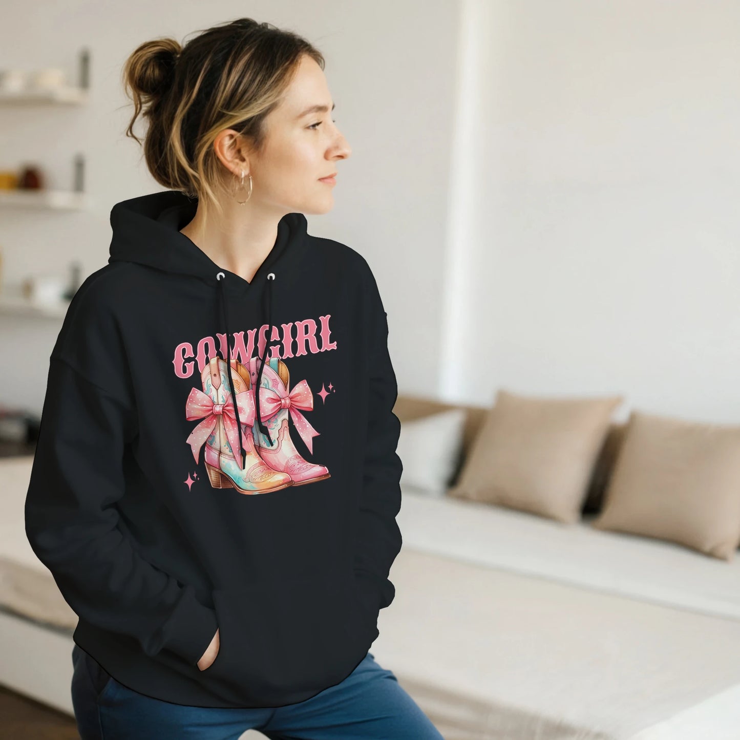 Woman wearing black cowgirl hoodie with pastel pink boots and bows graphic, front pouch pocket, drawstring hood.