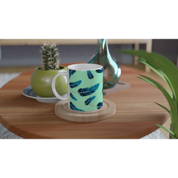 Blue Feather Mug in mint green with a blue feather pattern on a wooden table, surrounded by plants.