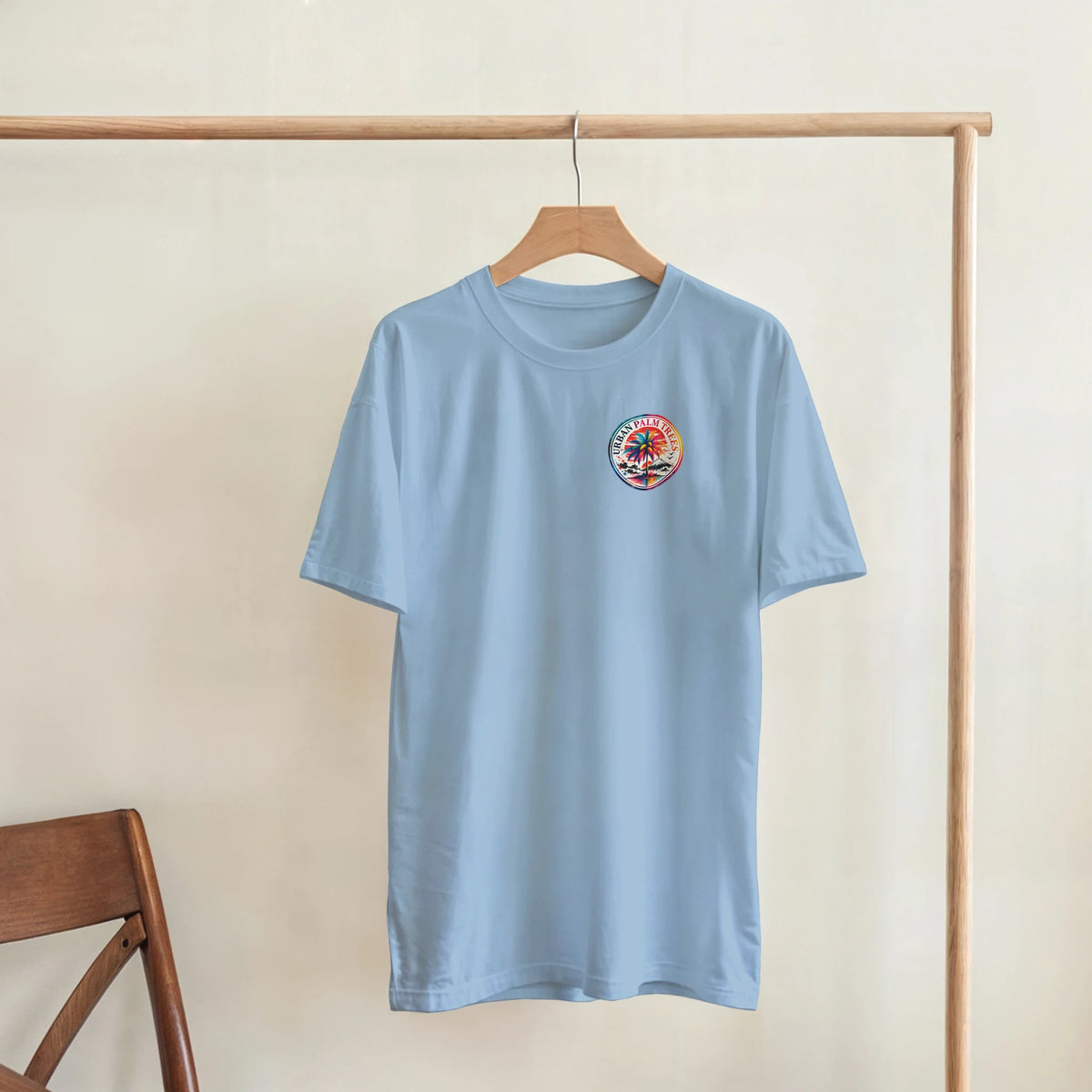 Light blue crew neck t-shirt featuring a retro Urban Palm Trees circular graphic on the chest, hanging on a wooden rack.