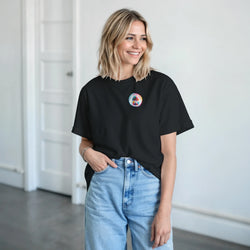 Woman wearing a black retro crew neck t-shirt with a small colorful circular palm tree graphic on the chest, paired with light blue jeans.